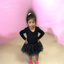Load image into Gallery viewer, Ek chhoti ladki, jiske baal safed ponytail band se bandhe hain, kala leotard aur sitaron se bhara kala tulle tutu pehne hue, kamar par haath rakhkar pose de rahi hai. Woh ek pink deewar ke samne khadi hai aur gulabi socks pehne hai.